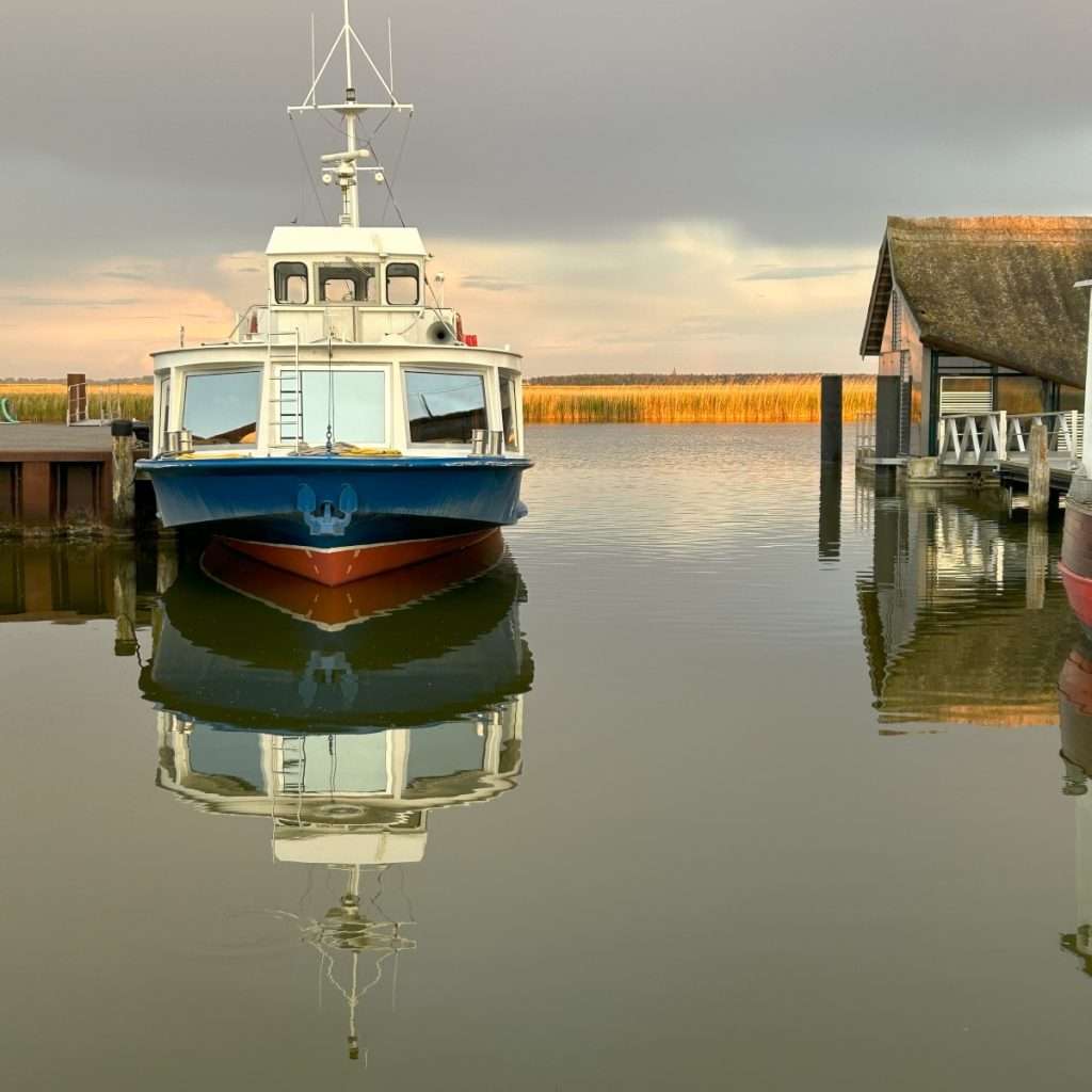 Fähre im Sonnenuntergang am Hafen von Zingst, dass sich im Wasser spiegelt. Rechts ist ein reetgedecktes Caféhaus zu sehen.