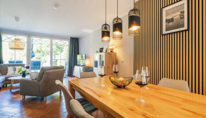 Essbereich für 6 Personen im Ferienhaus Herzmuschel in Zingst. Auf dem Holztisch stehen Weingläser. Die Wand ist mit modernen Akustikpaneelen verkleidet. Im Hintergrund sieht man den Wohnbereich.