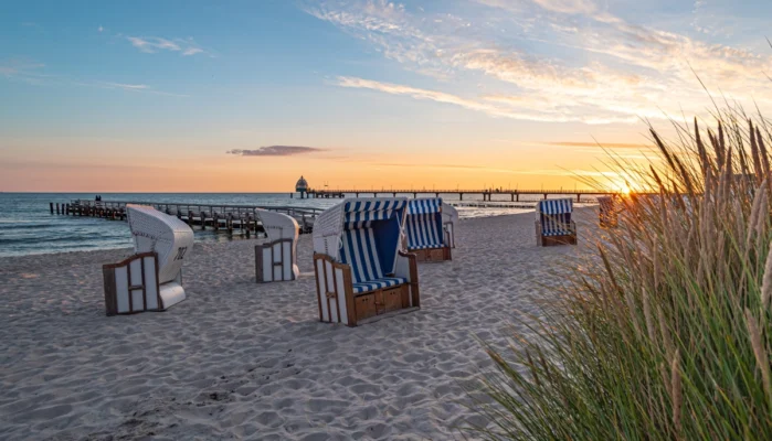 Strand von Zingst bei Sonnenaufgang. Im Vordergrund sind Strandkörbe zu sehen. Im Hintergrund sieht man die Seebrücke und Tauchglocke von Zingst.
